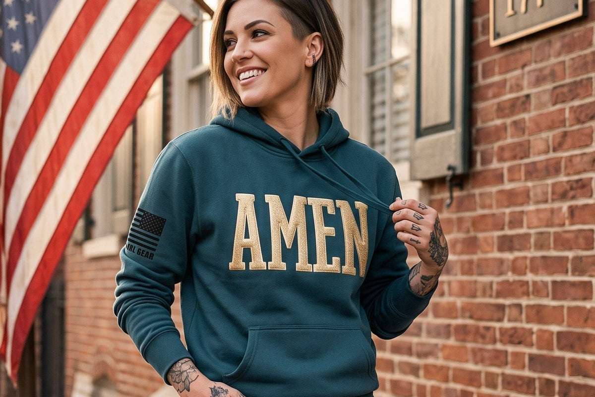 A woman wearing a teal hoodie with 'AMEN' printed in beige across the chest, paired with dark blue jeans, posing on the steps of a brick building with an American flag in the background.