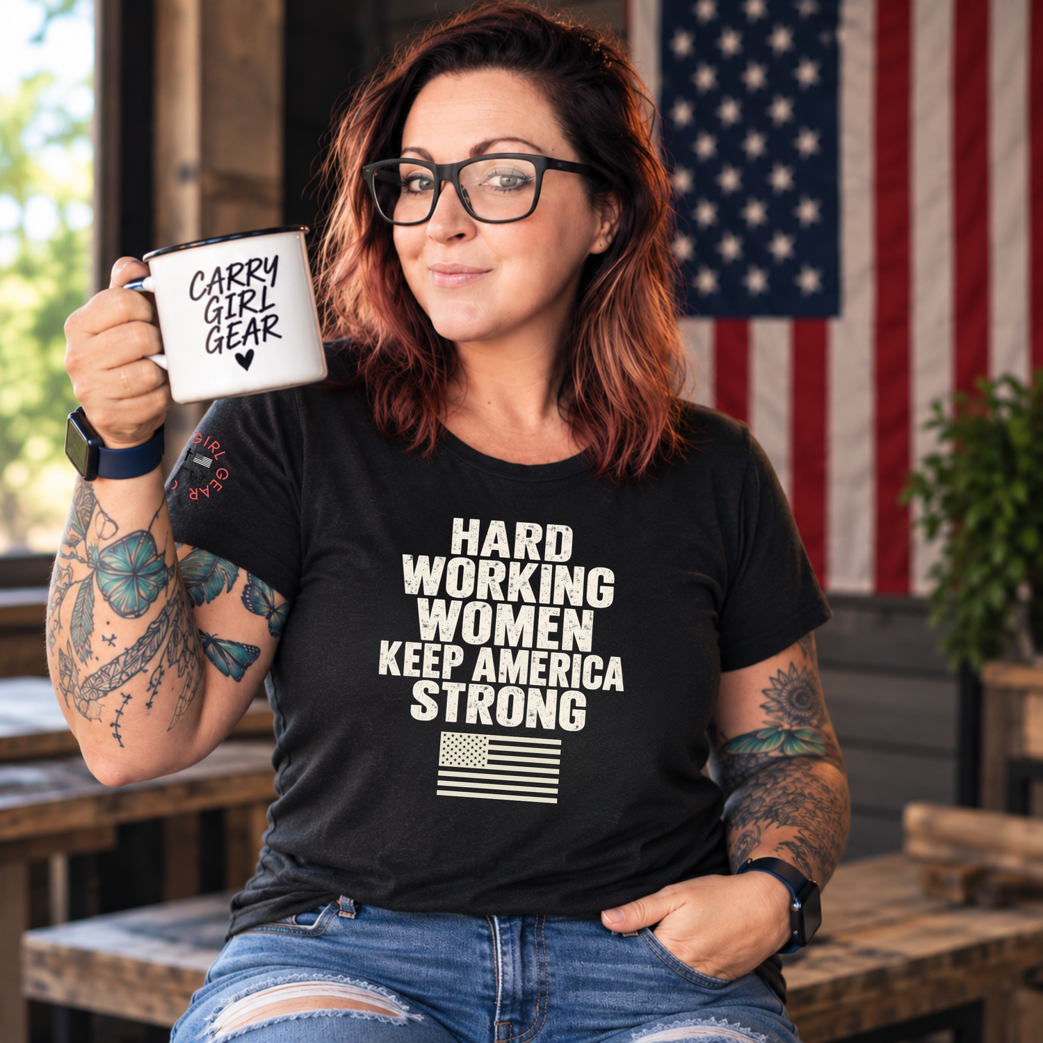 A woman with glasses and tattoos holds a white mug that reads 'CARRY GIRL GEAR' while wearing a black graphic T-shirt with the text 'HARD WORKING WOMEN KEEP AMERICA STRONG' and a small American flag graphic. The background shows an American flag and rustic wooden furniture.