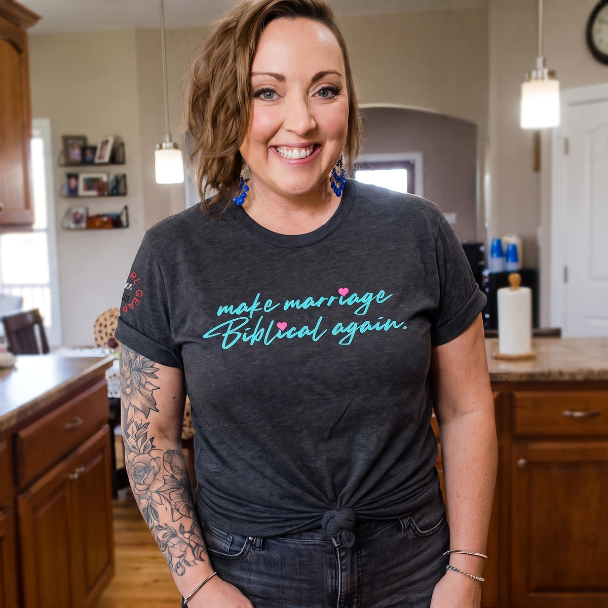 A smiling woman with short brown hair and a tattooed arm stands in a kitchen wearing a dark charcoal gray t-shirt with the phrase 'make marriage Biblical again.' printed in light blue script with a small pink heart accent.