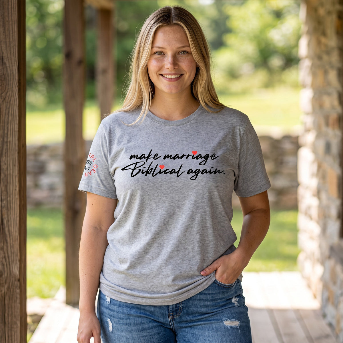 A woman wearing a heather gray graphic t-shirt with the text 'make marriage Biblical again.' printed in black script with small red heart accents, standing on a wooden porch with a blurred outdoor background.