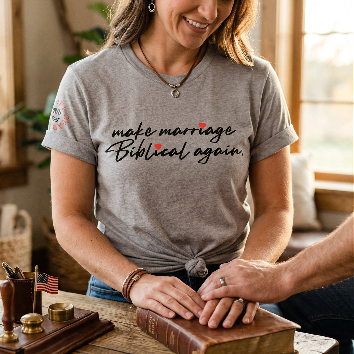 A woman wearing a gray graphic t-shirt with the text 'make marriage Biblical again.' in black script with small red heart accents, sitting at a wooden table with a man's hands resting on an old leather-bound Bible.