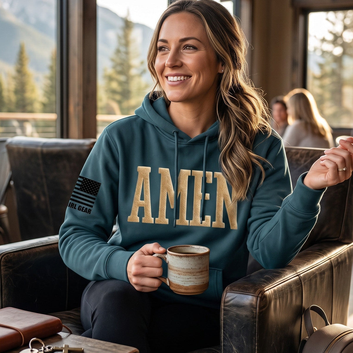 A woman sitting in a rustic cafe chair wearing a teal hoodie with 'AMEN' printed in gold across the chest, holding a ceramic mug. The background shows a cozy cafe interior with mountain scenery visible through large windows.