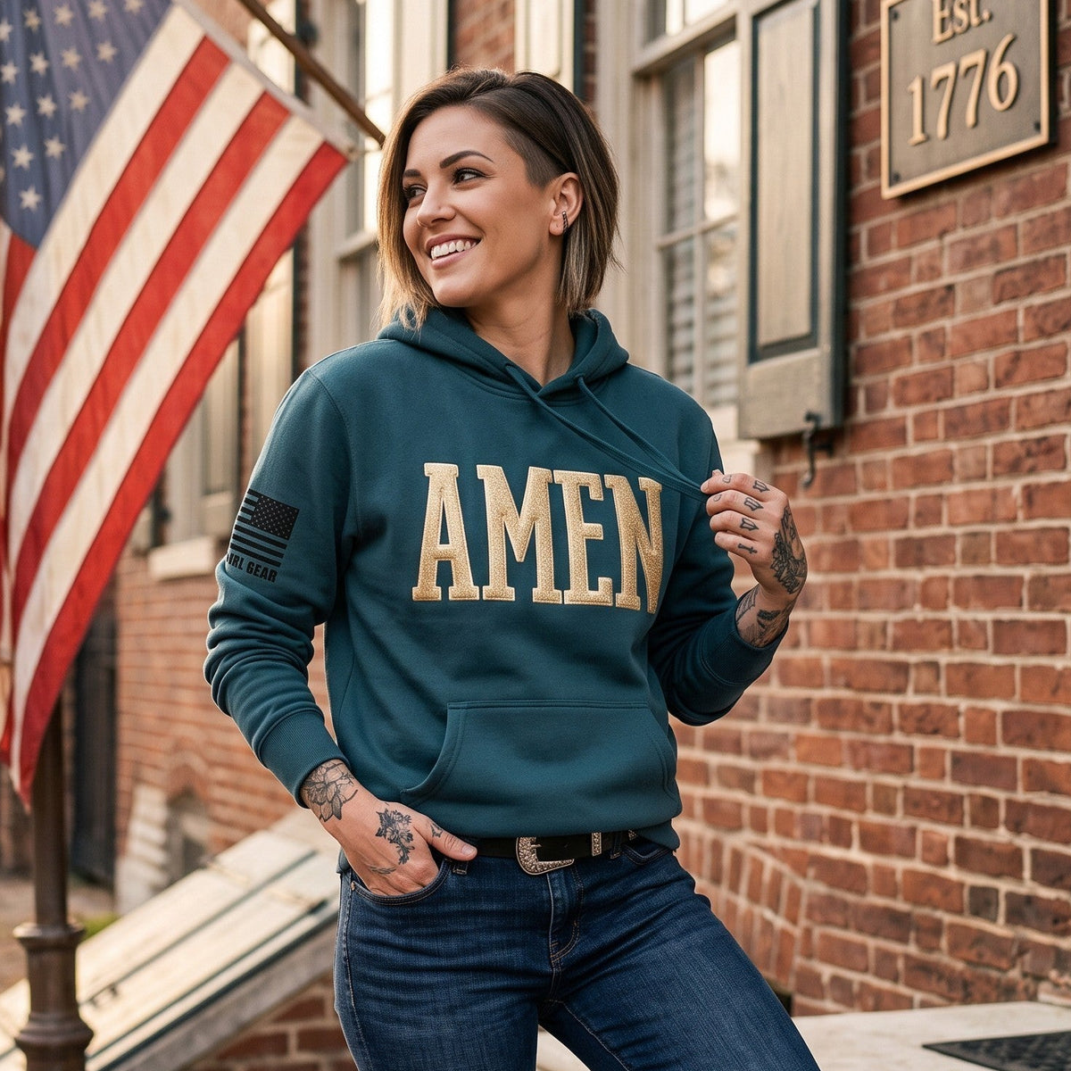 A woman wearing a teal hoodie with 'AMEN' printed in beige across the chest, paired with dark blue jeans, posing on the steps of a brick building with an American flag in the background.