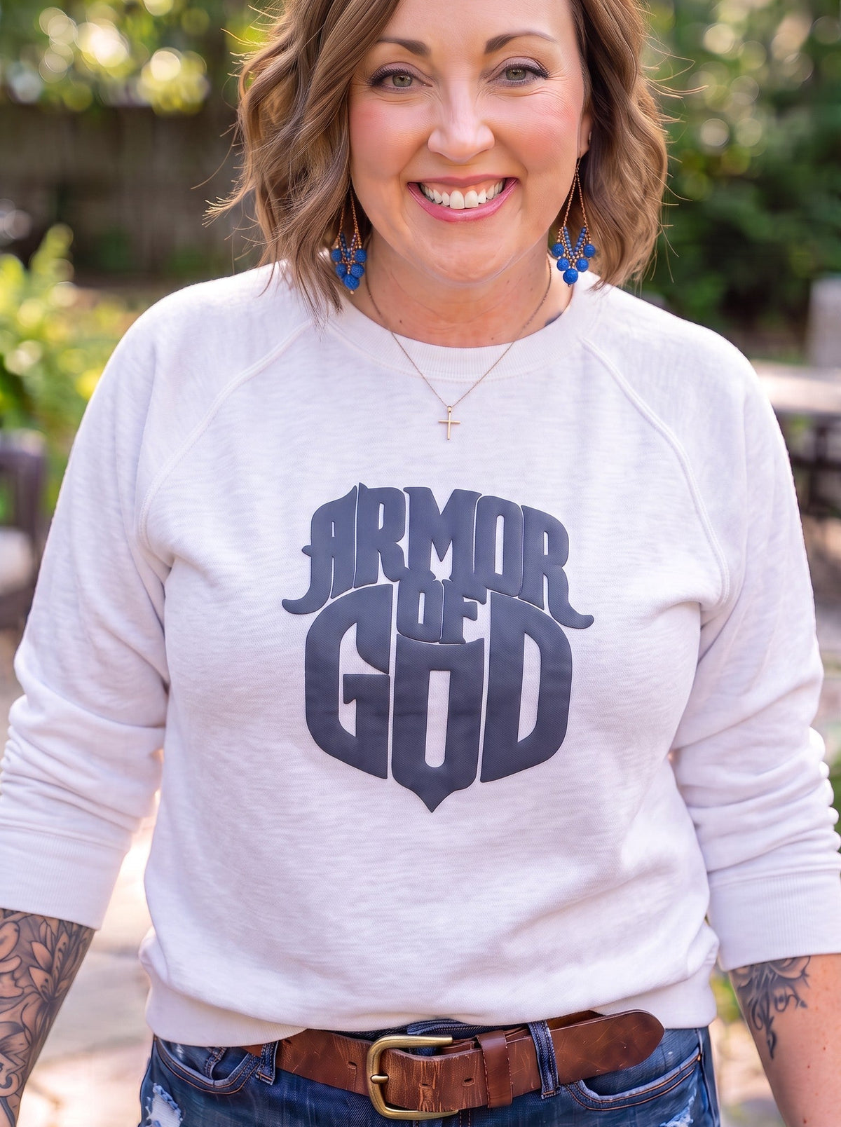A smiling woman wearing a white long-sleeve crewneck sweatshirt with a dark navy graphic that reads 'ARMOR OF GOD' on the front, paired with blue jeans and a brown leather belt, standing outdoors in front of a blurred green background.