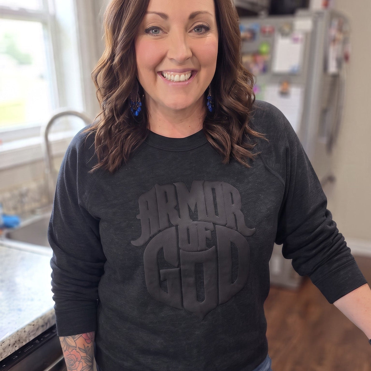 A woman standing in a kitchen wearing a dark charcoal gray long-sleeve crewneck sweatshirt with a tonal 'ARMOR OF GOD' graphic on the front, paired with blue jeans and black boots.