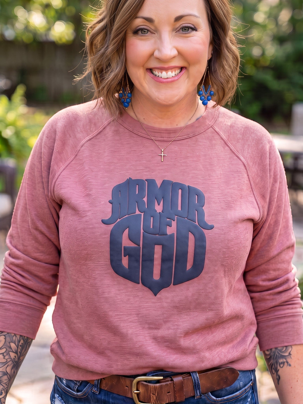 A smiling woman wearing a dusty rose long-sleeve crewneck sweatshirt with a dark navy graphic that reads 'ARMOR OF GOD' on the front, paired with blue jeans and a brown belt, standing outdoors in front of a blurred green garden background.