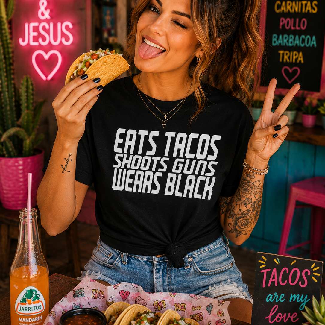 A woman in a black graphic t-shirt with white text reading 'EATS TACOS SHOOTS GUNS WEARS BLACK' poses in a colorful restaurant setting, holding a taco and making a peace sign. A table in front of her has tacos, a bottle of hot sauce, and a drink.