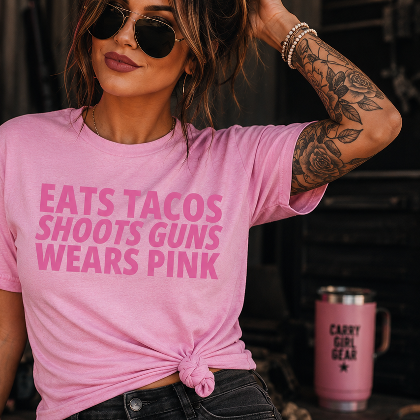 A woman wearing a pink graphic t-shirt with the text 'EATS TACOS SHOOTS GUNS WEARS PINK' in pink, paired with distressed black jeans. She is posing in a rustic setting with a pink neon sign that reads 'Faith Family Freedom' in the background.