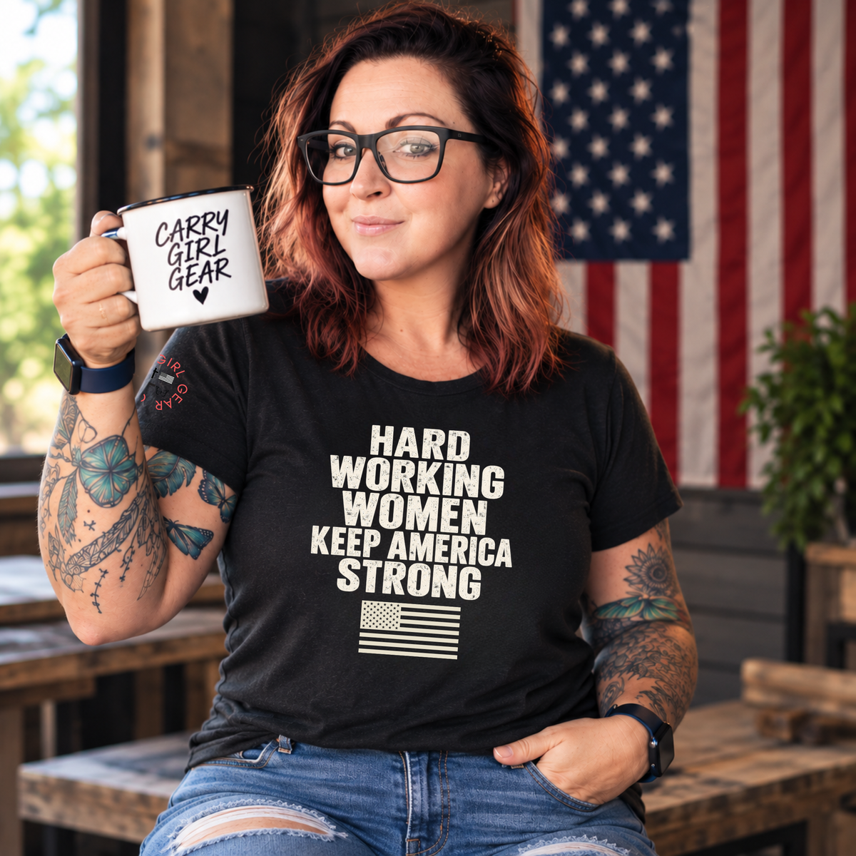 A woman with glasses and tattoos holds a white mug that reads 'CARRY GIRL GEAR' while wearing a black graphic T-shirt with the text 'HARD WORKING WOMEN KEEP AMERICA STRONG' and a small American flag graphic. The background shows an American flag and rustic wooden furniture.