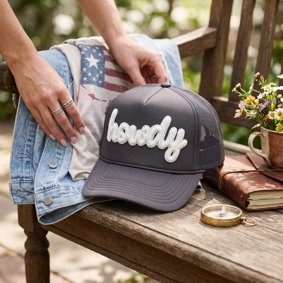 A dark gray trucker hat with white cursive embroidery reading 'howdy' sits on a rustic wooden bench. A person's hands are visible adjusting the brim, with a denim jacket, t-shirt, flowers, a compass, and a leather-bound book arranged around it in an outdoor garden setting.