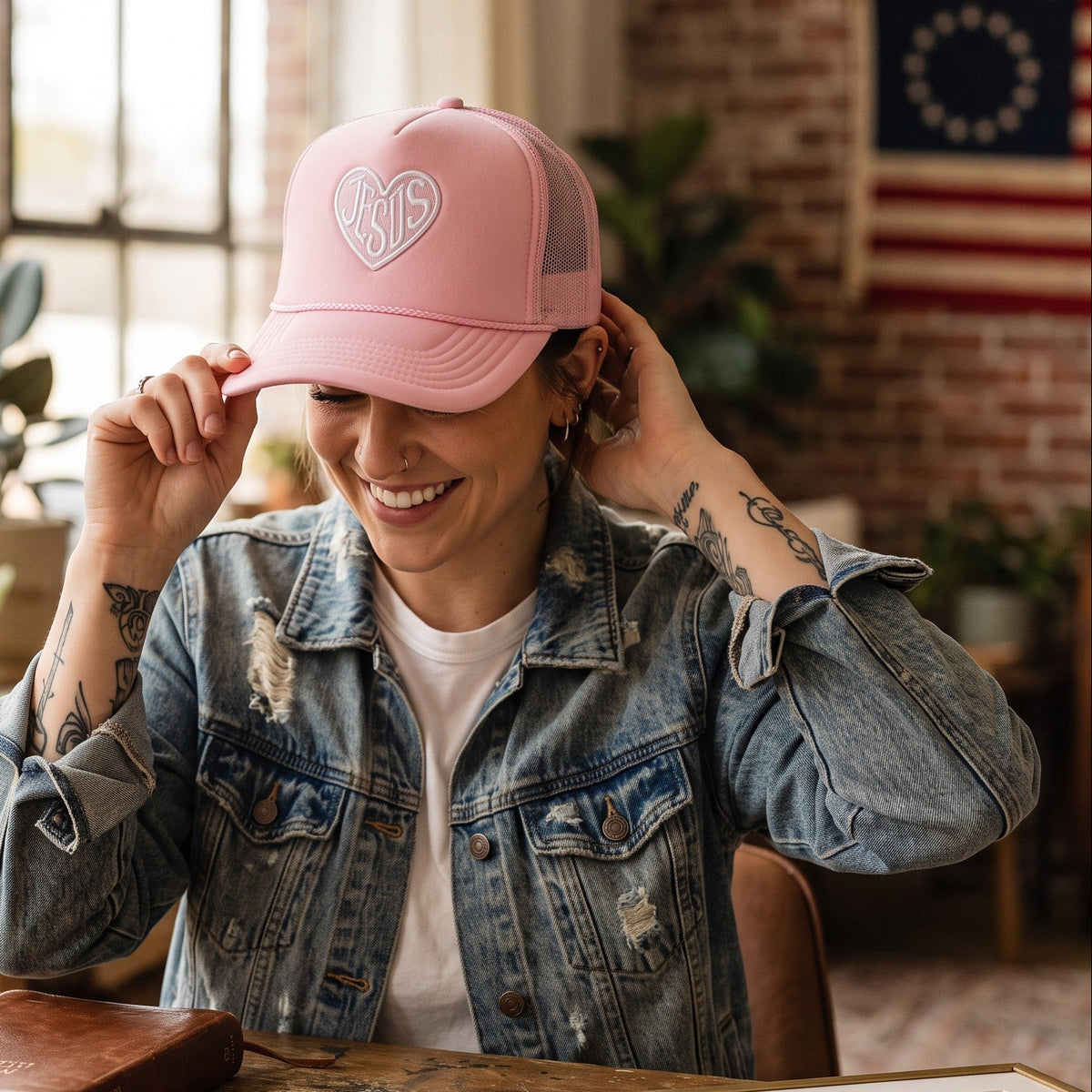 A woman in a rustic studio setting wearing a pink trucker hat with a white embroidered heart and the words 'FAT HAPPY'. She is wearing a distressed denim jacket over a white t-shirt, sitting at a wooden desk with art supplies, a mug, and framed prints.