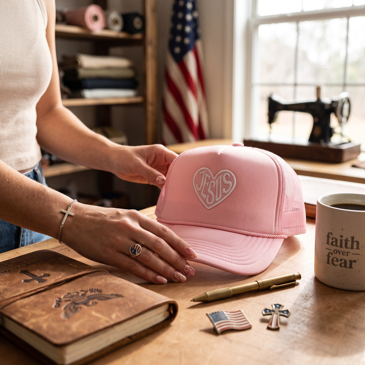 A person in a white sleeveless top holds a light pink trucker cap with a white embroidered 'Jesus' heart logo on a wooden table. The scene includes a brown leather journal, a pen, a small American flag pin, a cross pin, and a coffee mug with the text 'faith over fear' in a warm, rustic shop setting.