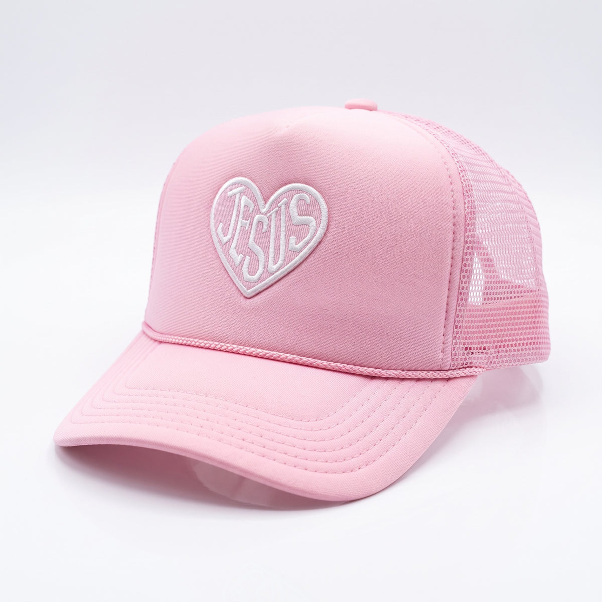 A light pink trucker-style cap with a curved brim, featuring a white embroidered heart outline on the front containing the word 'JESUS' in white script.