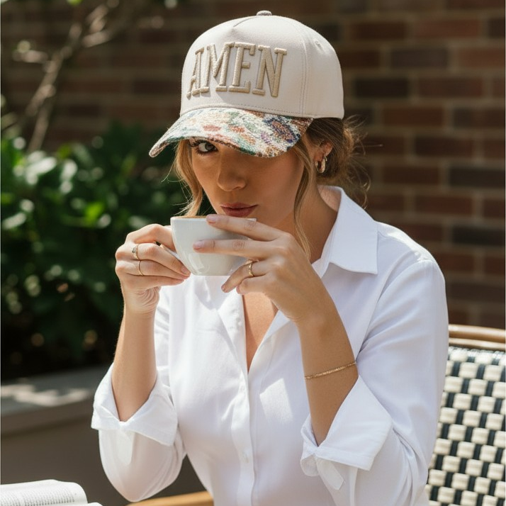 Woman sitting outdoors, wearing a cap with 'AMEN' text, holding a cup.