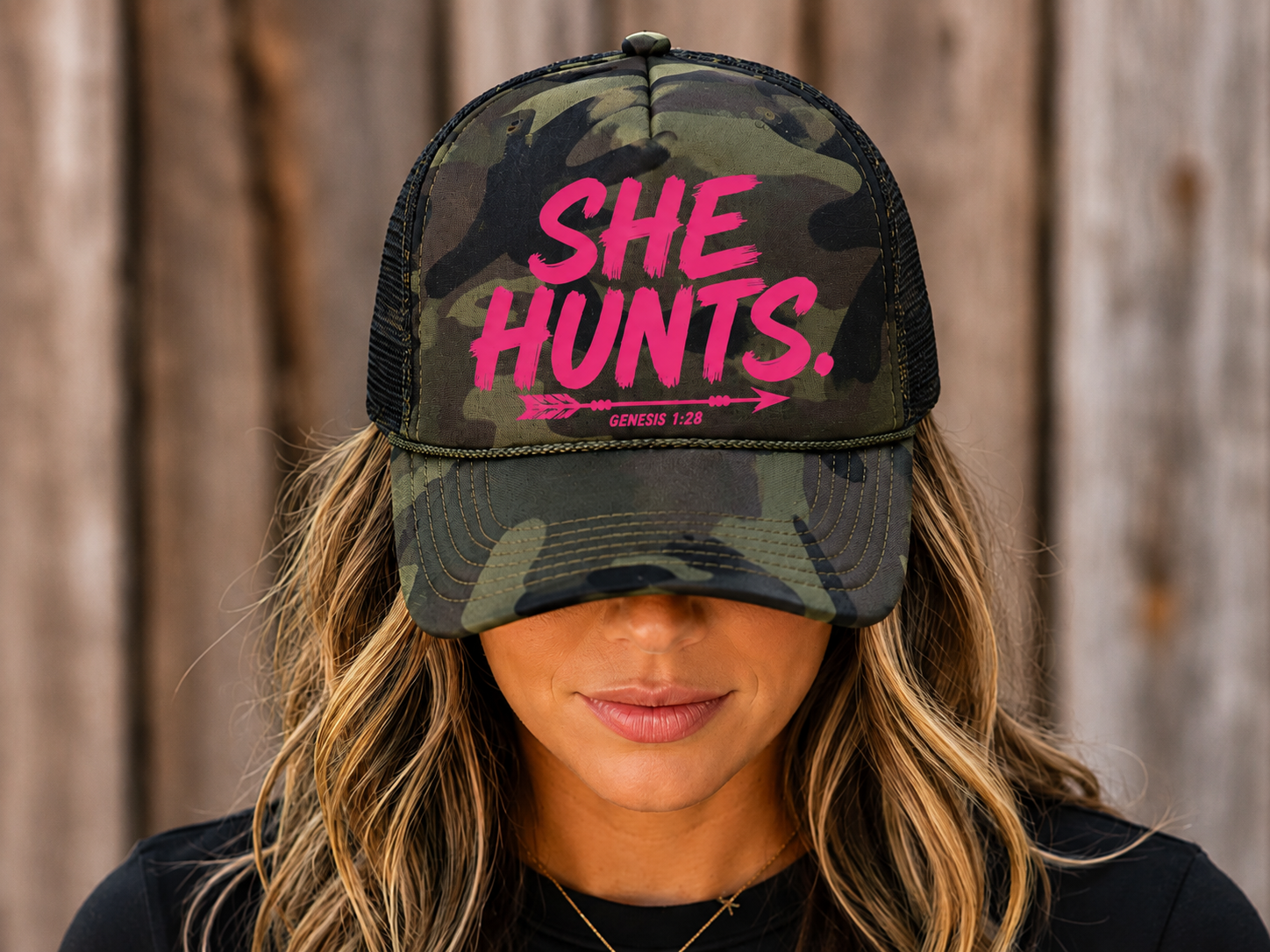 A woman wearing a camouflage trucker hat with pink text that reads 'SHE HUNTS.' and smaller text 'GENESIS 1:28' underneath, photographed against a rustic wooden background.