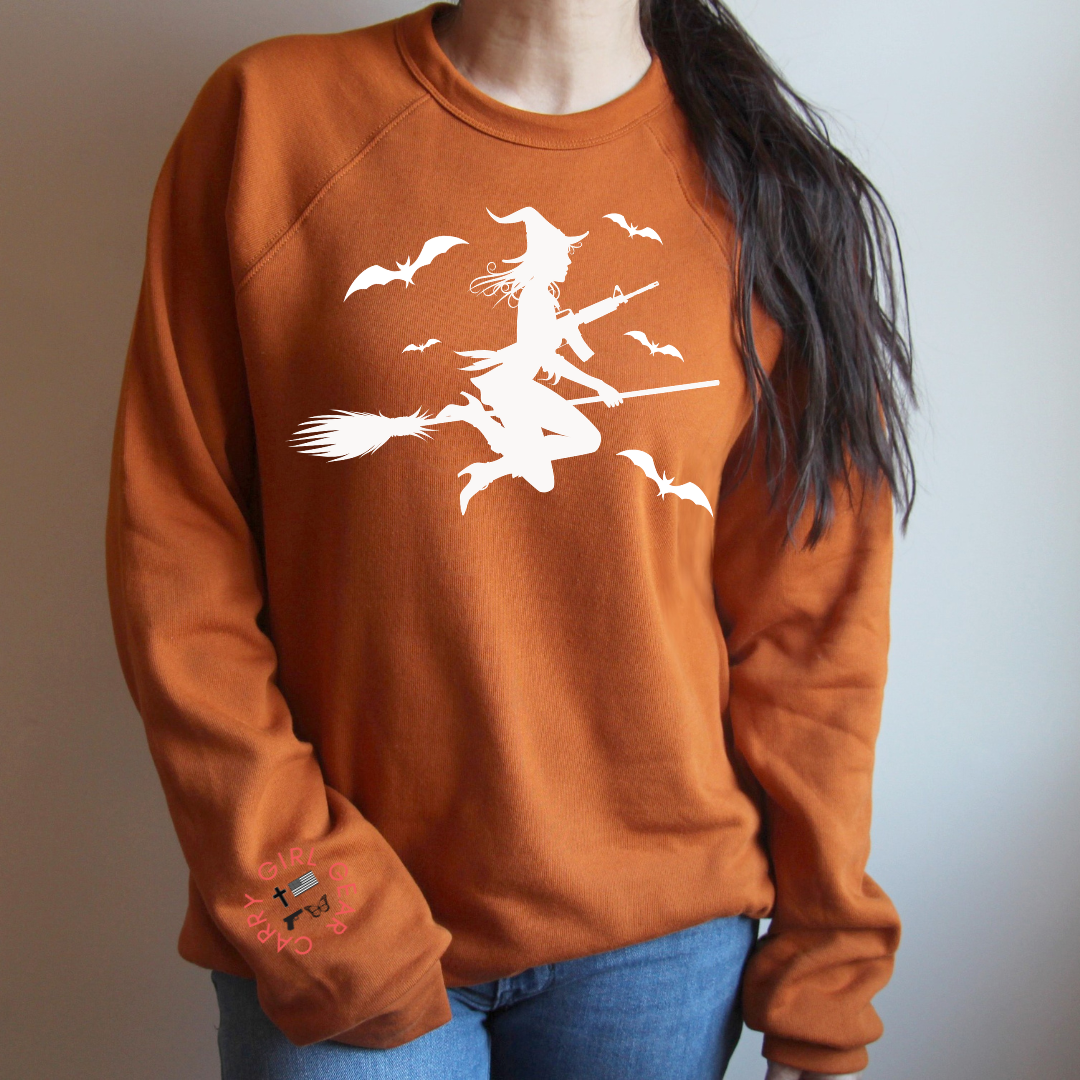 A woman wearing a rust-orange crewneck sweatshirt with a white graphic of a witch riding a broomstick surrounded by bats. The sweatshirt has a small embroidered design on the left sleeve.