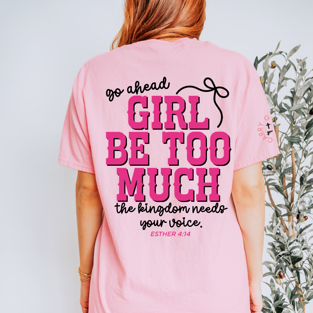 A woman shown from the back wearing a light pink graphic T-shirt with bold text reading 'go ahead GIRL BE TOO MUCH the kingdom needs your voice. ESTHER 4:14'.
