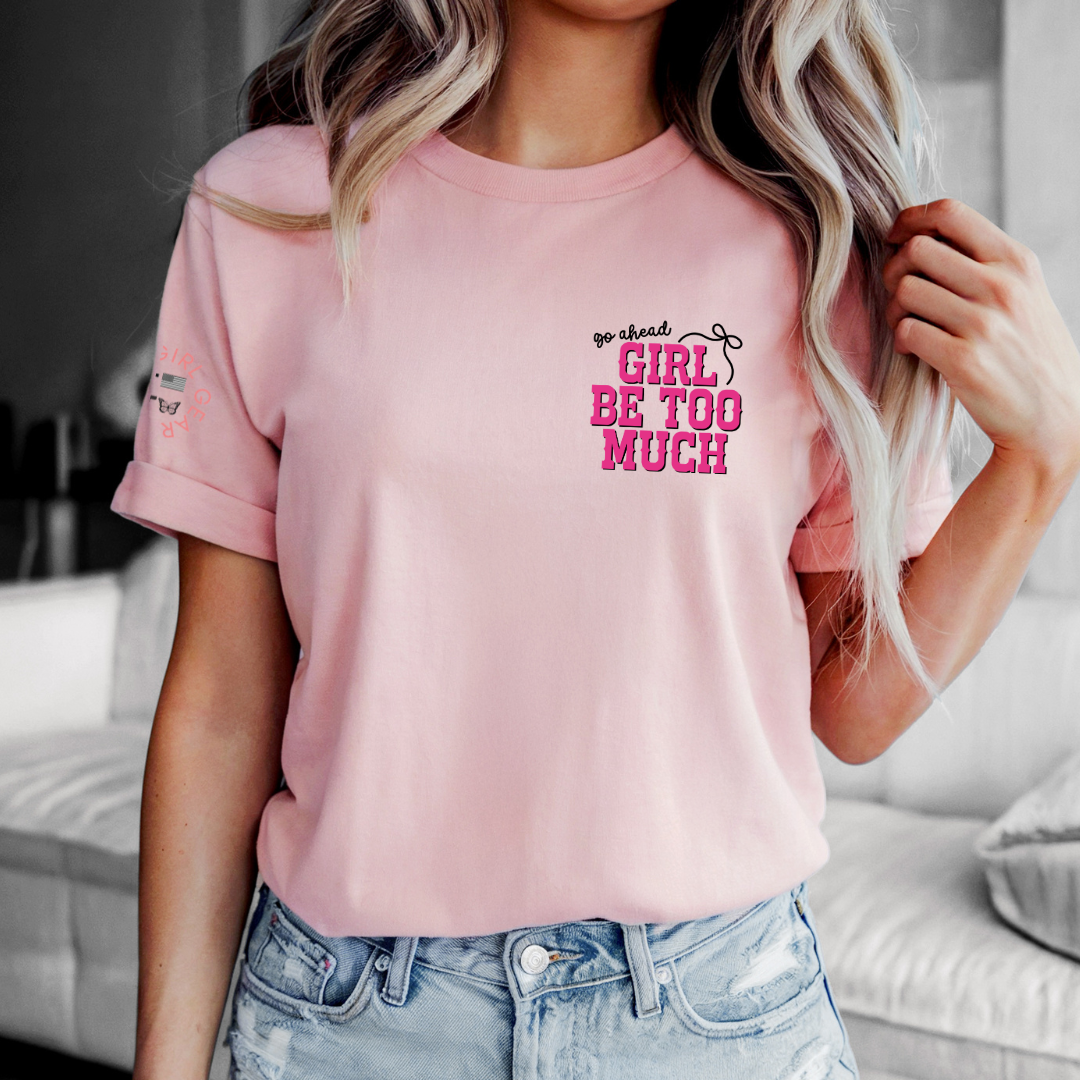 A woman wearing a light pink crewneck t-shirt with a graphic that reads 'go ahead GIRL BE TOO MUCH' in pink and black text, paired with light-wash blue jeans in a modern living room setting.