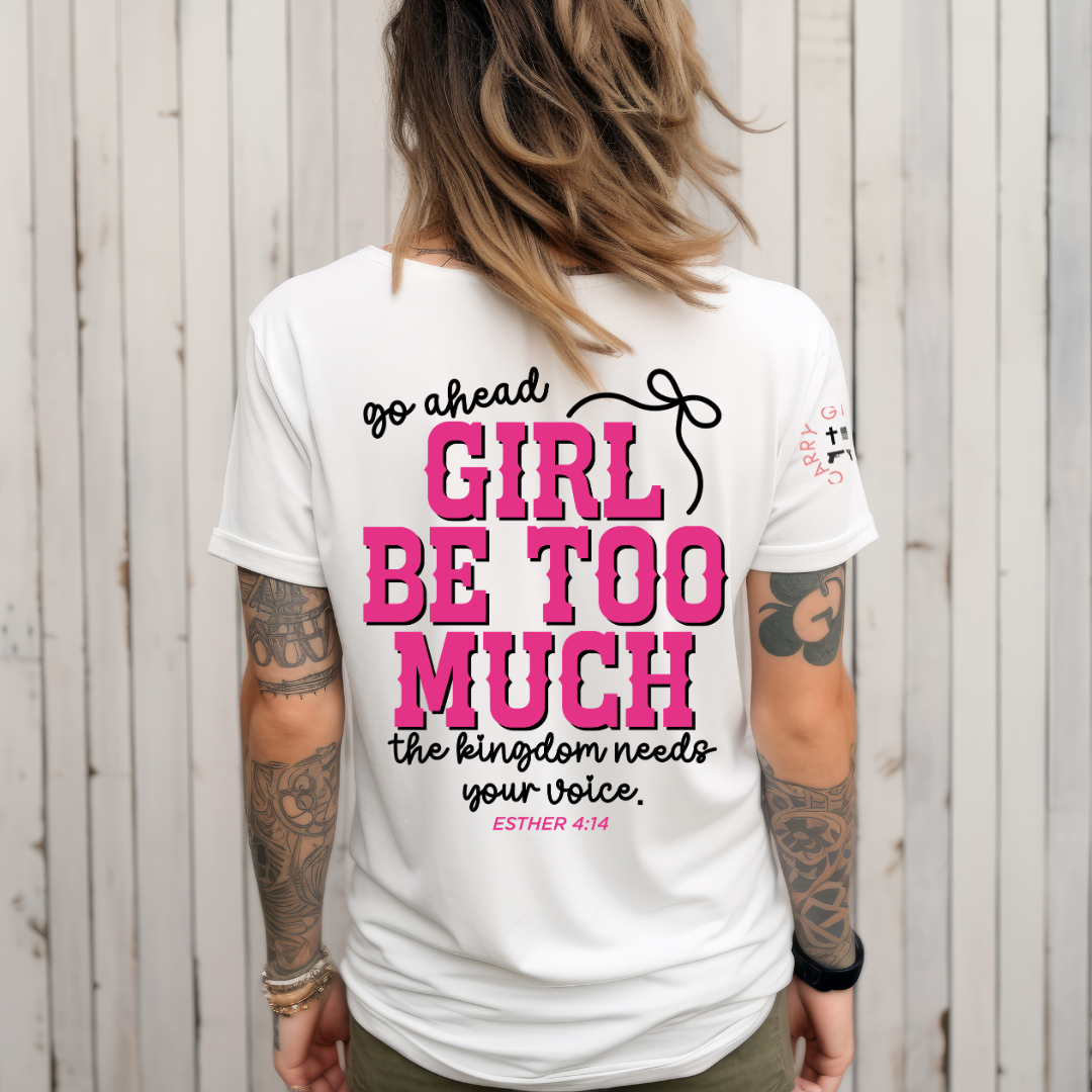 A woman viewed from behind wearing a white graphic T-shirt with bold pink and black text reading 'go ahead GIRL BE TOO MUCH the kingdom needs your voice. ESTHER 4:14'.