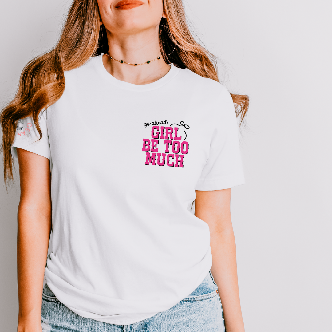A woman wearing a white short-sleeve graphic T-shirt with the text 'go ahead GIRL BE TOO MUCH' in pink and black, paired with light blue jeans against a light gray background.