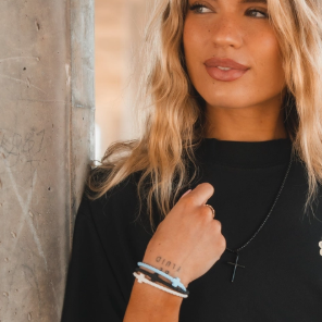 Woman wearing a black top and multiple bracelets, standing against a concrete wall.
