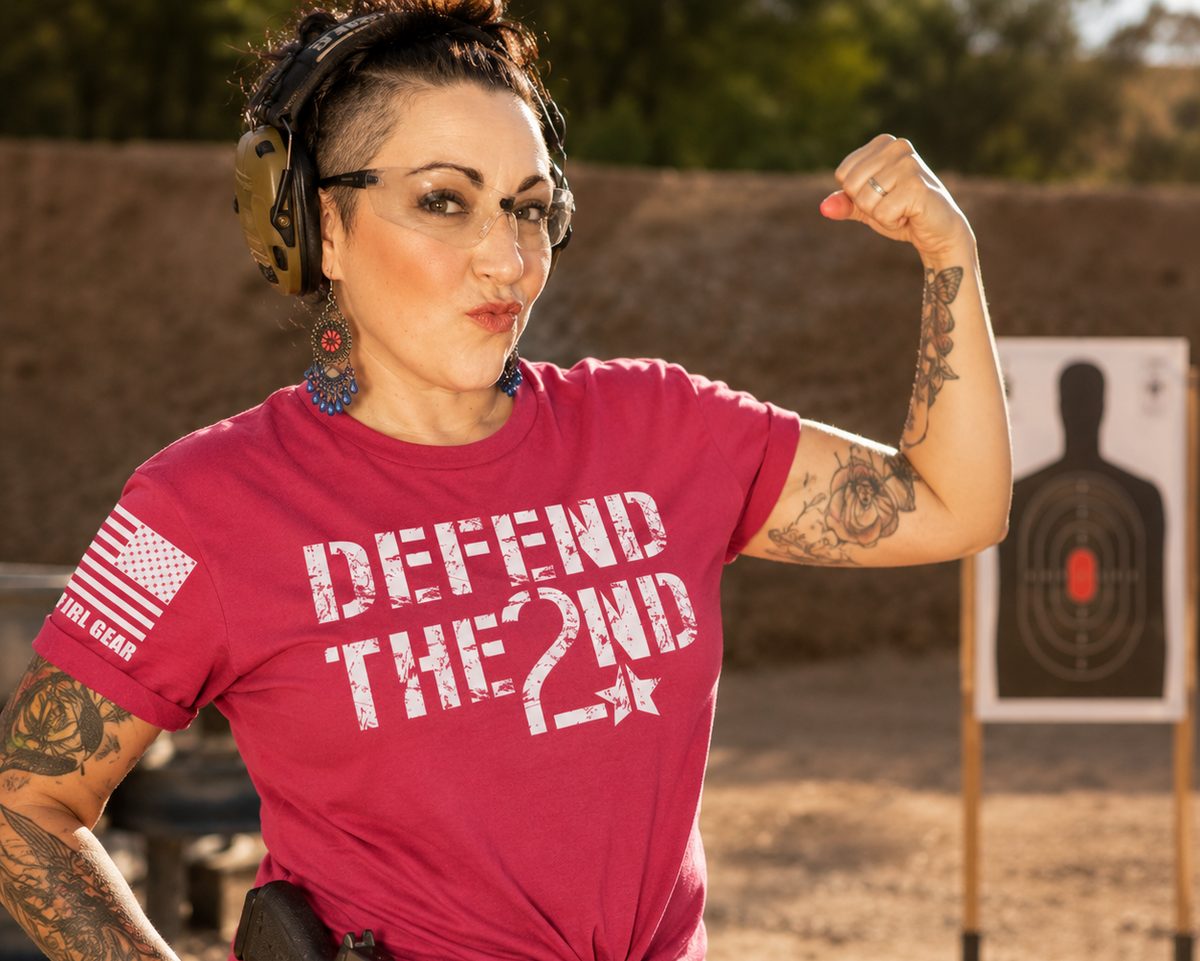A woman at an outdoor shooting range wearing a bright pink t-shirt with 'DEFEND THE 2ND' printed in white distressed text, paired with a black belt and holster, posing with a raised fist toward a target backdrop.