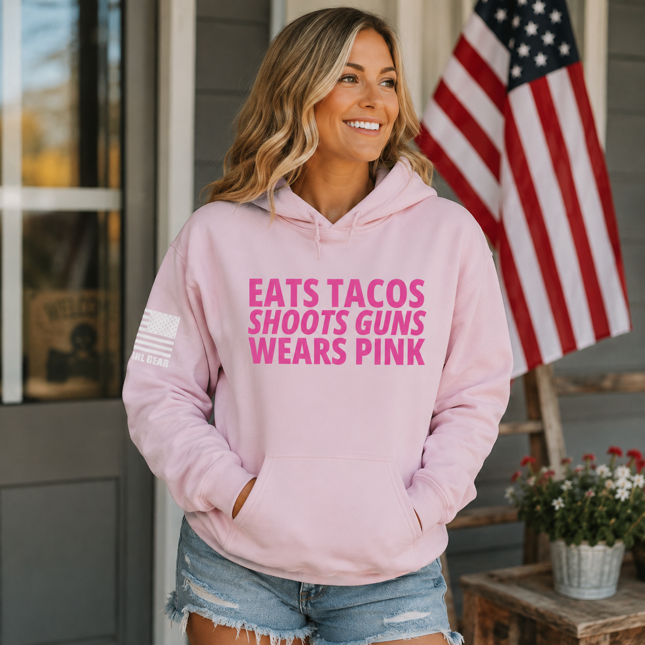 A woman wearing a light pink hoodie with bold pink text reading 'EATS TACOS SHOOTS GUNS WEARS PINK' and a small American flag graphic on the sleeve, standing on a porch with an American flag in the background.