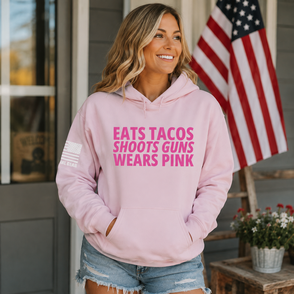 A woman wearing a light pink hoodie with bold pink text reading 'EATS TACOS SHOOTS GUNS WEARS PINK' and a small American flag graphic on the sleeve, standing on a porch with an American flag in the background.