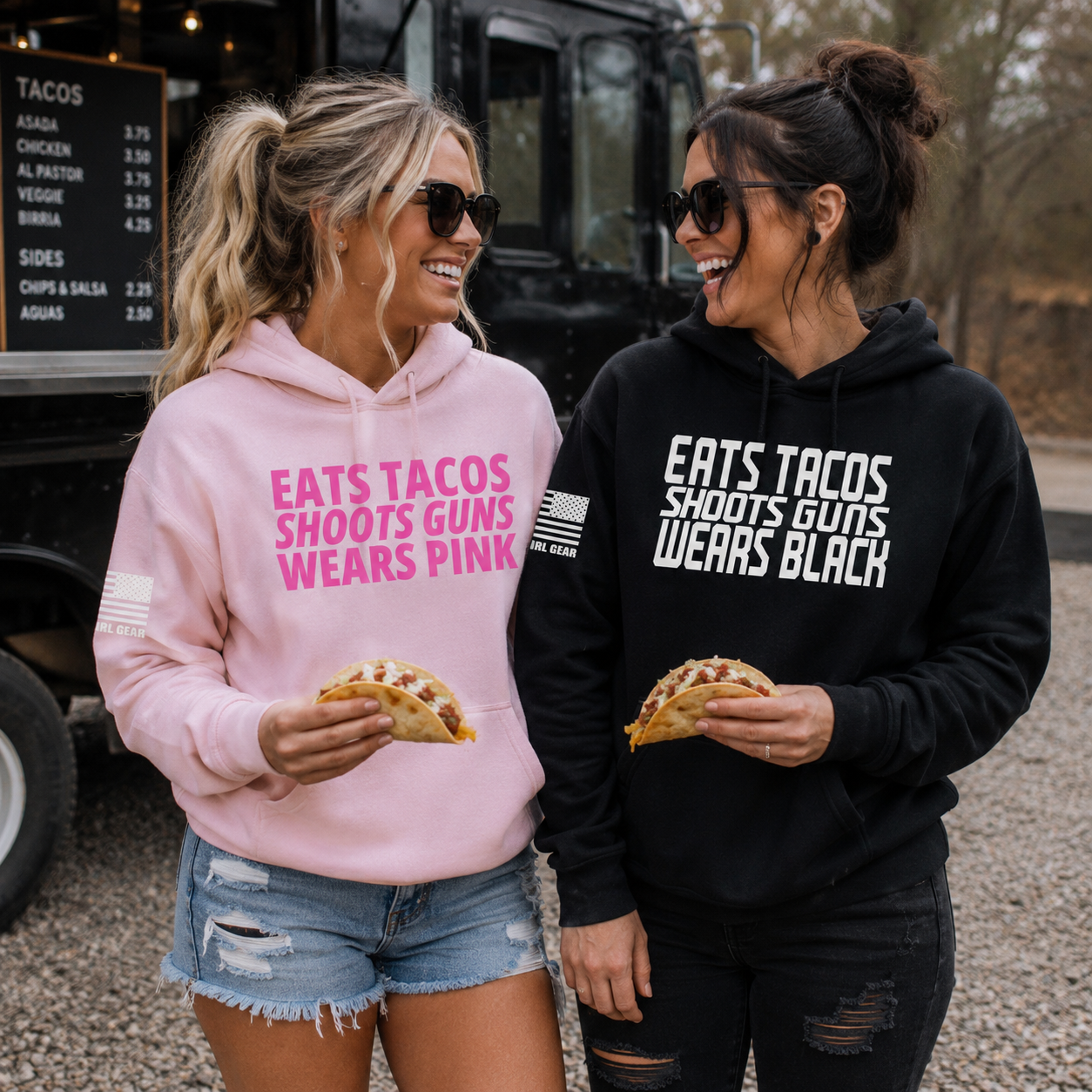 Two women standing outdoors in front of a food truck, each wearing a graphic hoodie. The woman on the left wears a light pink hoodie with bright pink text reading 'EATS TACOS SHOOTS GUNS WEARS PINK' and denim shorts.
