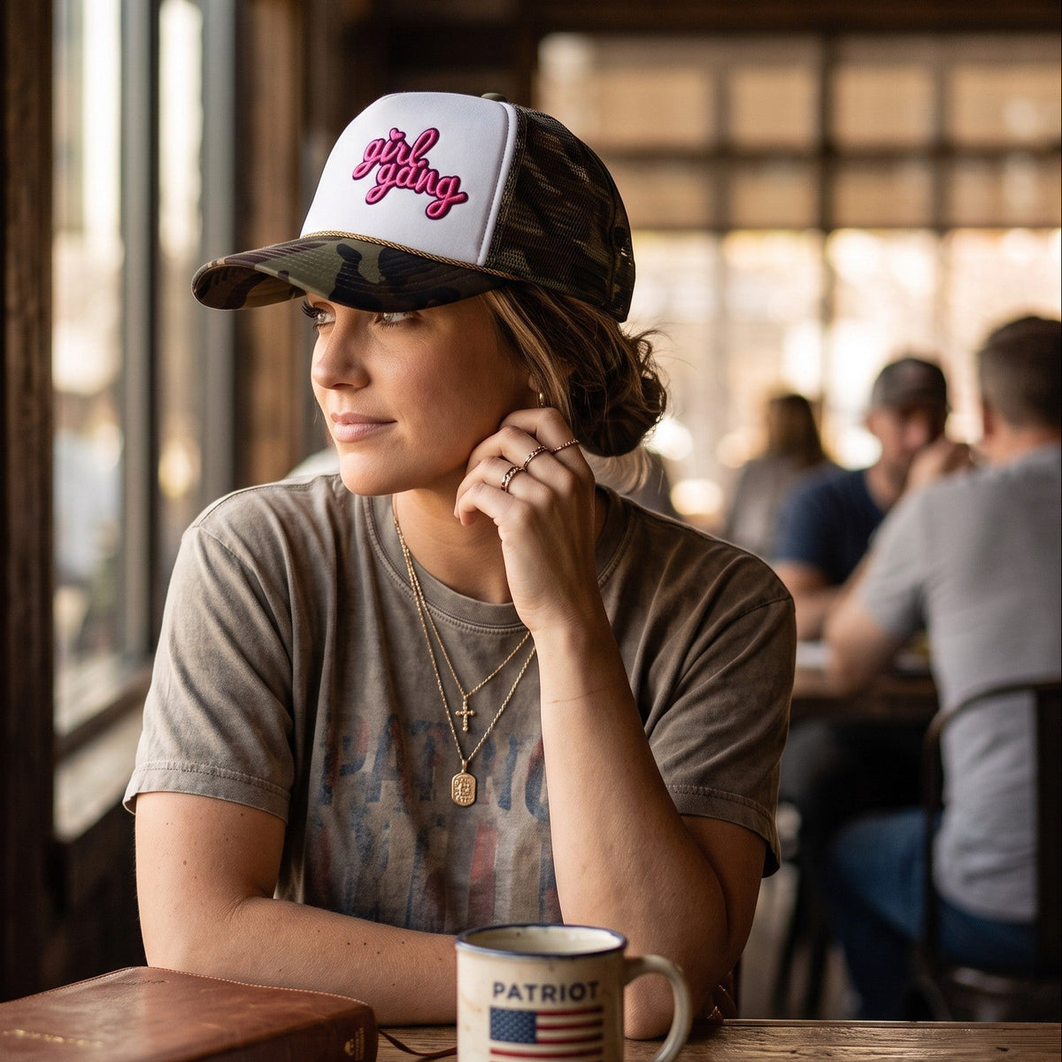 A woman sits at a wooden table in a rustic cafe, wearing a camouflage and white trucker hat with the brand name 'Patriot' embroidered in red on the front. A matching white Patriot mug sits on the table in front of her, alongside a vintage book and eyeglasses.