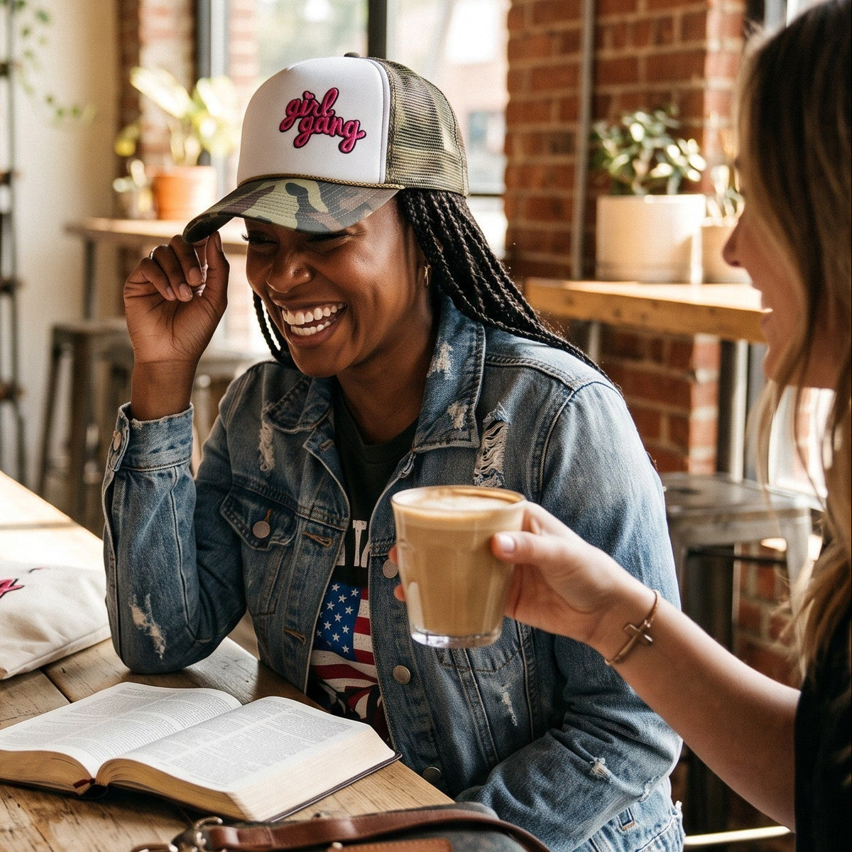 Three women laughing together at a rustic wooden cafe table. The central woman wears a camouflage and white trucker hat with a pink script logo, a distressed denim jacket, and a graphic t-shirt.