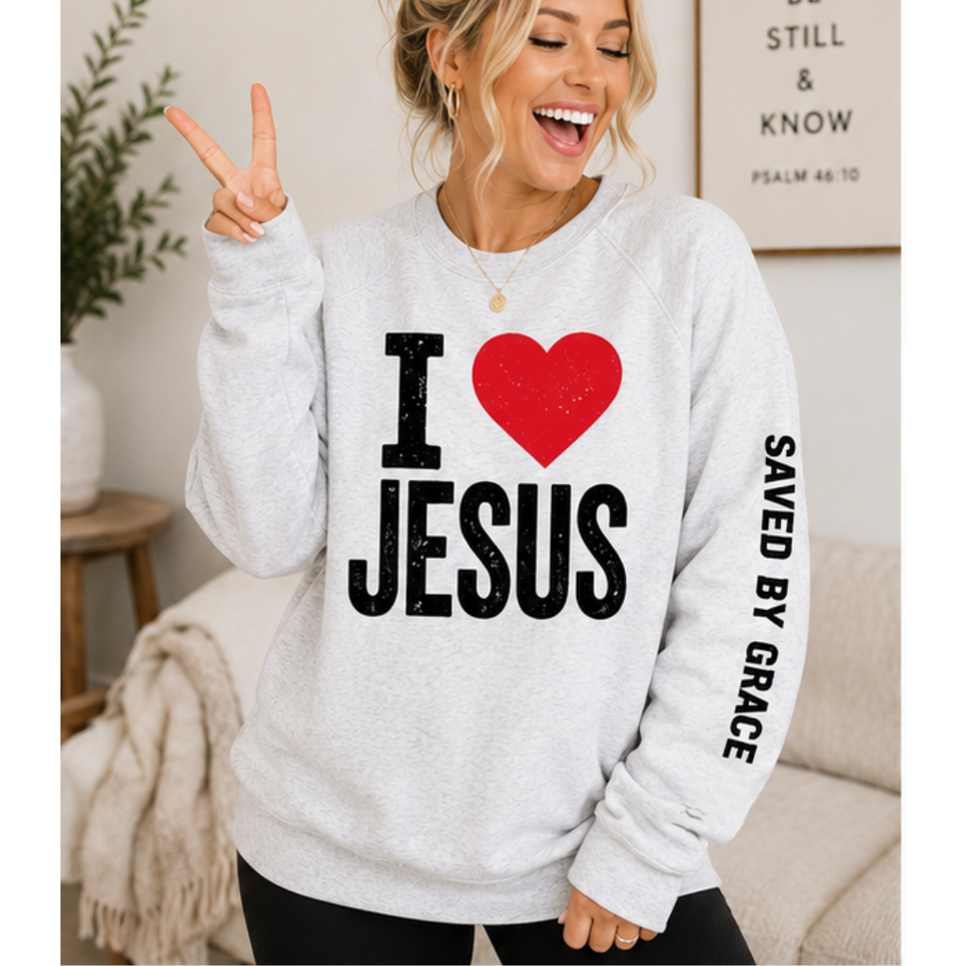 A woman wearing a light heather gray crewneck sweatshirt with a graphic that reads 'I [red heart] JESUS' and 'SAVED BY GRACE' printed on the sleeve, posing in a cozy living room setting.