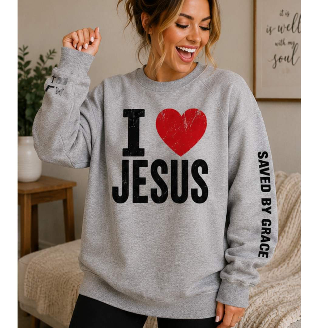 A woman wearing a heather gray crewneck sweatshirt with a graphic that reads 'I [red heart] JESUS' and 'SAVED BY GRACE' on the sleeve, posed in a cozy indoor setting.