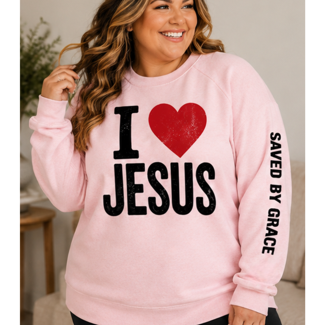 A smiling woman wearing a light pink crewneck sweatshirt with a bold graphic on the front reading 'I [heart] JESUS' in black and red, and 'SAVED BY GRACE' printed vertically in black on the right sleeve.