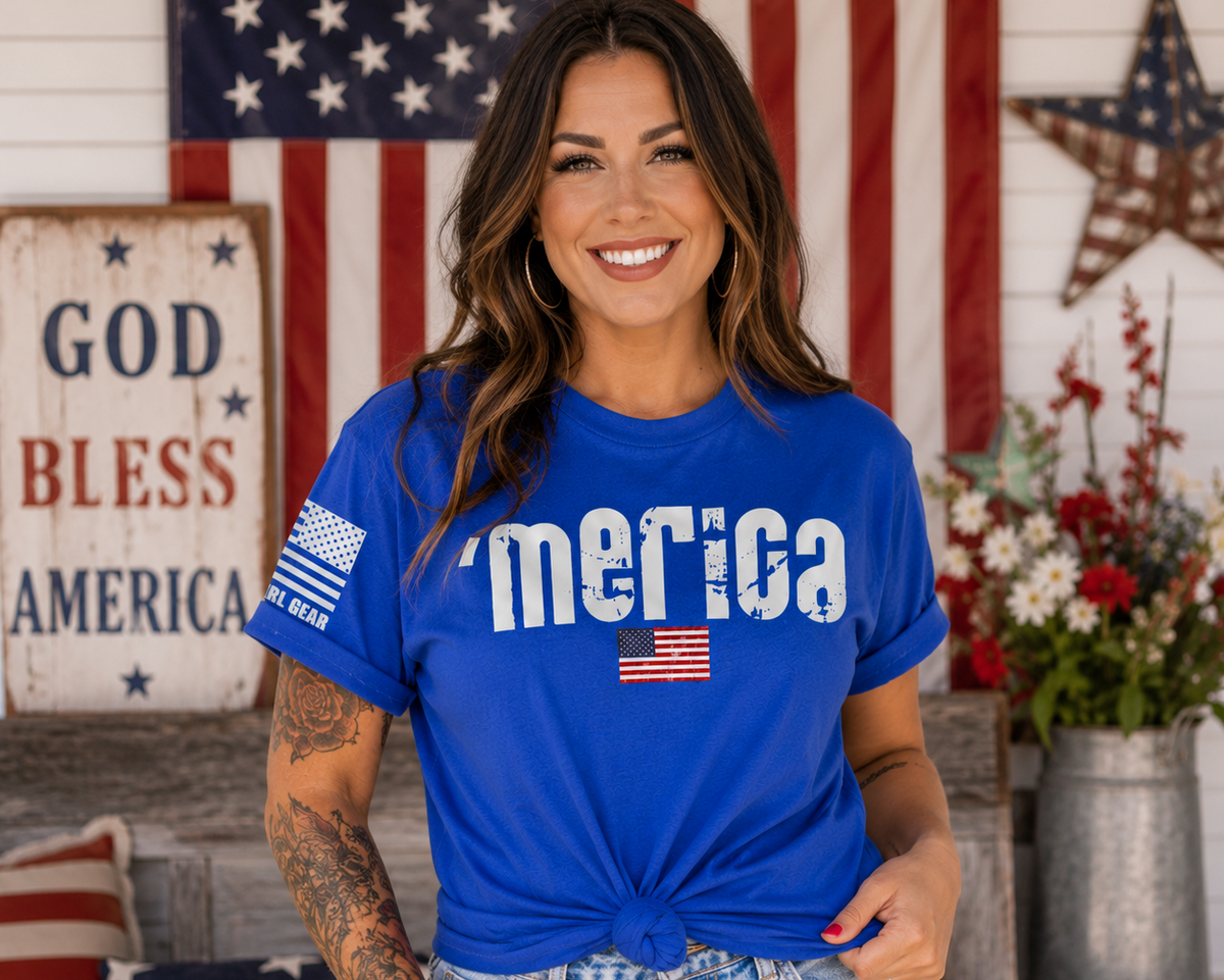 A woman wearing a bright blue t-shirt with 'America' printed in distressed white letters and small American flag graphics on the sleeve and chest, posed in front of a rustic patriotic background with an American flag and a 'God Bless America' sign.