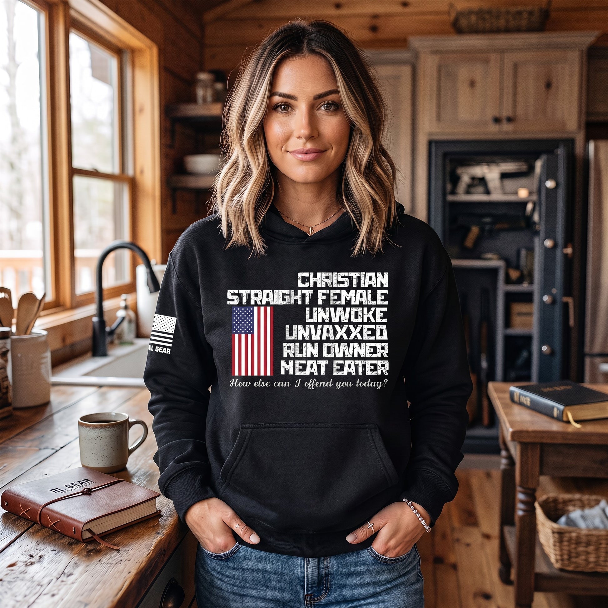 A woman wearing a black graphic hoodie stands in a rustic wooden kitchen. The hoodie features a white text graphic that reads 'CHRISTIAN STRAIGHT FEMALE UNWOXED UNVAXXED RUN OWNER MEAT EATER' with a small American flag graphic and a smaller white text line underneath.