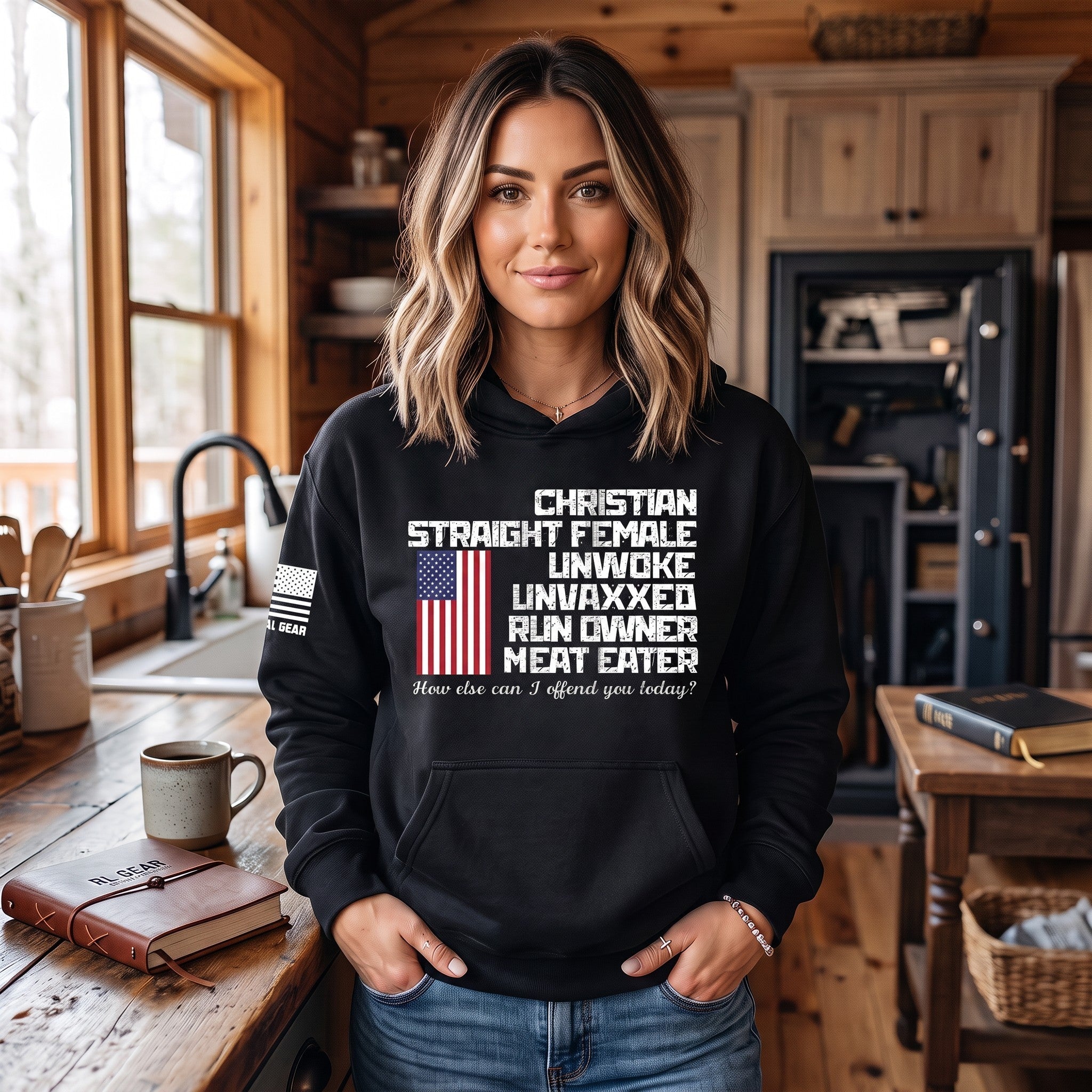 A woman wearing a black graphic hoodie stands in a rustic wooden kitchen. The hoodie features a white text graphic that reads 'CHRISTIAN STRAIGHT FEMALE UNWOXED UNVAXXED RUN OWNER MEAT EATER' with a small American flag graphic and a smaller white text line underneath.