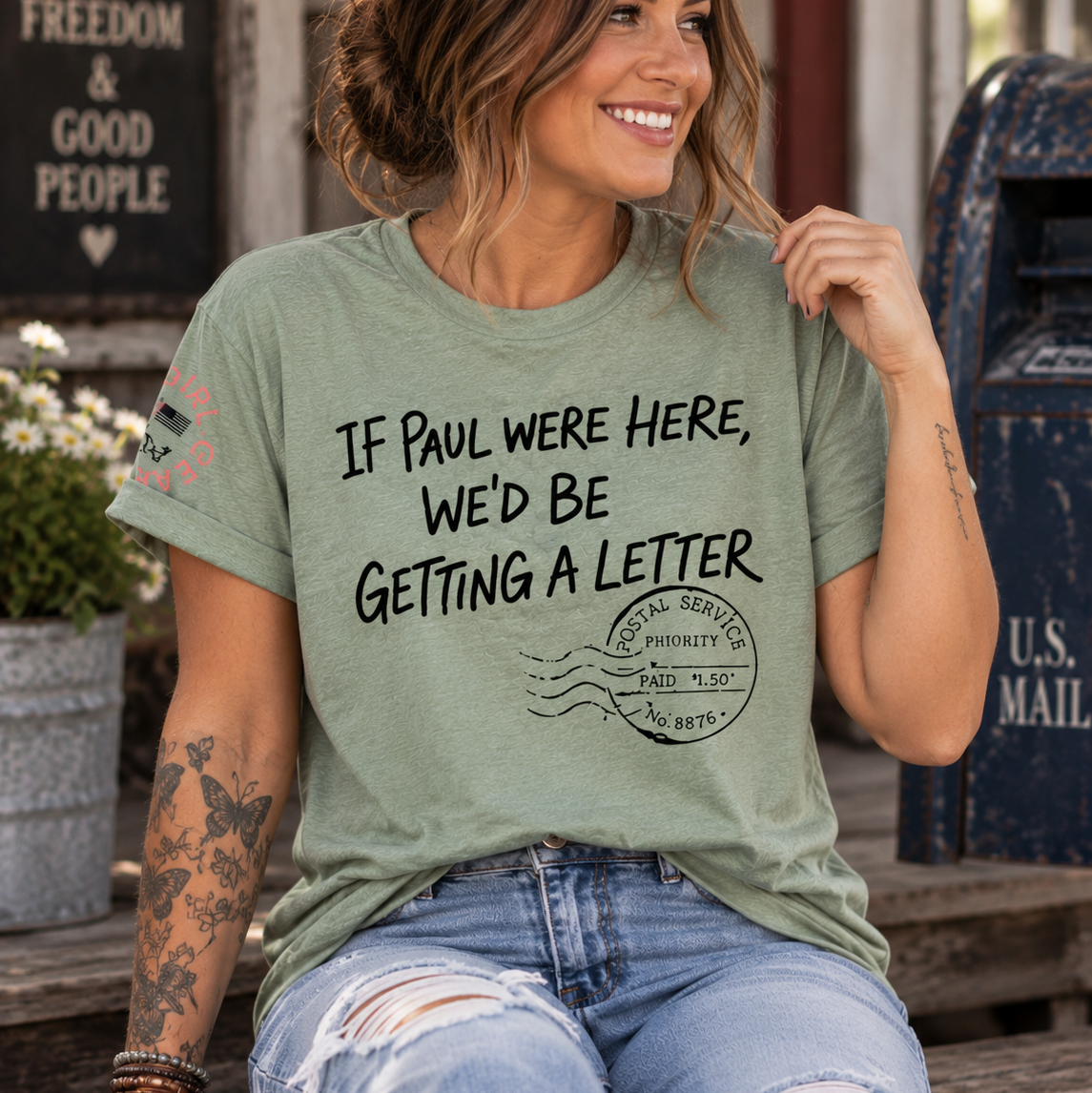 A woman wearing a light sage green graphic t-shirt with a black printed message about a letter, paired with distressed blue jeans, sitting outdoors in front of a rustic wooden door and a blue mailbox.