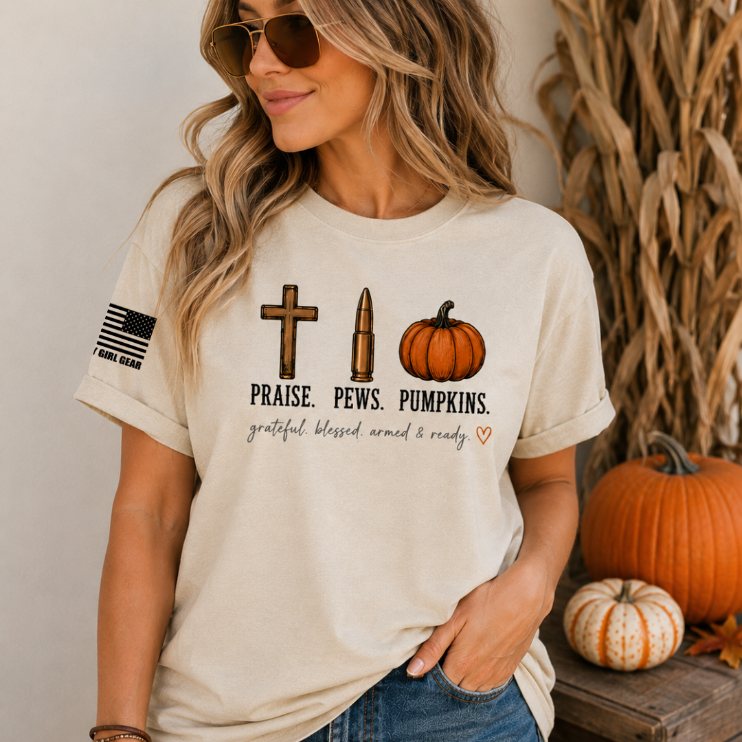 A woman wearing a beige graphic t-shirt with a fall harvest theme, featuring a cross, bullet, and pumpkin graphic with the text 'PRAISE. PEWS.