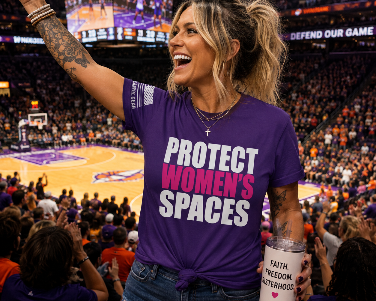 A woman cheering at a WNBA basketball game wearing a purple graphic t-shirt that reads 'PROTECT WOMEN'S SPACES' in white and pink text, holding a white tumbler with 'FAITH, FREEDOM, SISTERHOOD' printed on it.