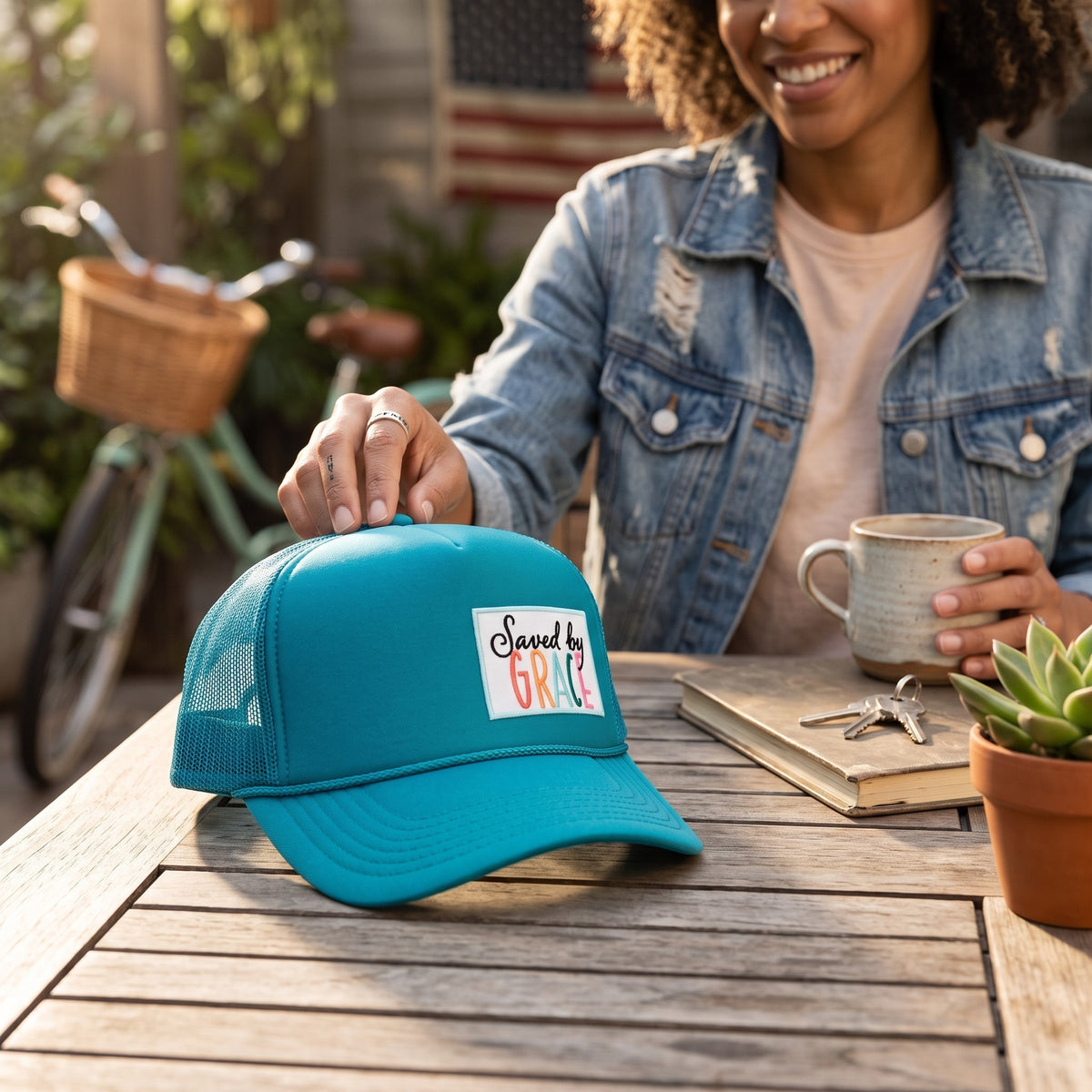 A woman in a denim jacket sits at a wooden table holding a mug, while a turquoise trucker hat with a 'Saved by GARY' patch rests on the table in front of her. The setting is a sunny, outdoor patio with a bicycle and plants in the background.