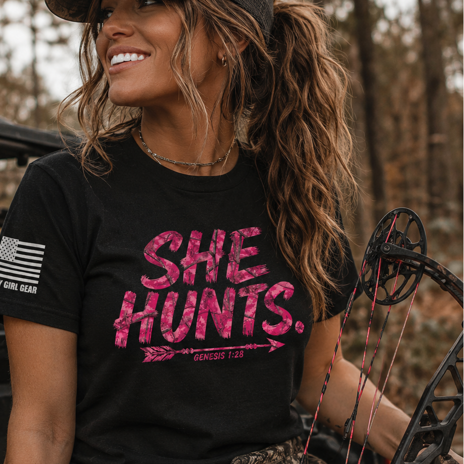 A woman in camouflage clothing and a camo cap sits in the bed of a pickup truck, holding a compound bow. She is wearing a black graphic t-shirt with the text 'SHE HUNTS.