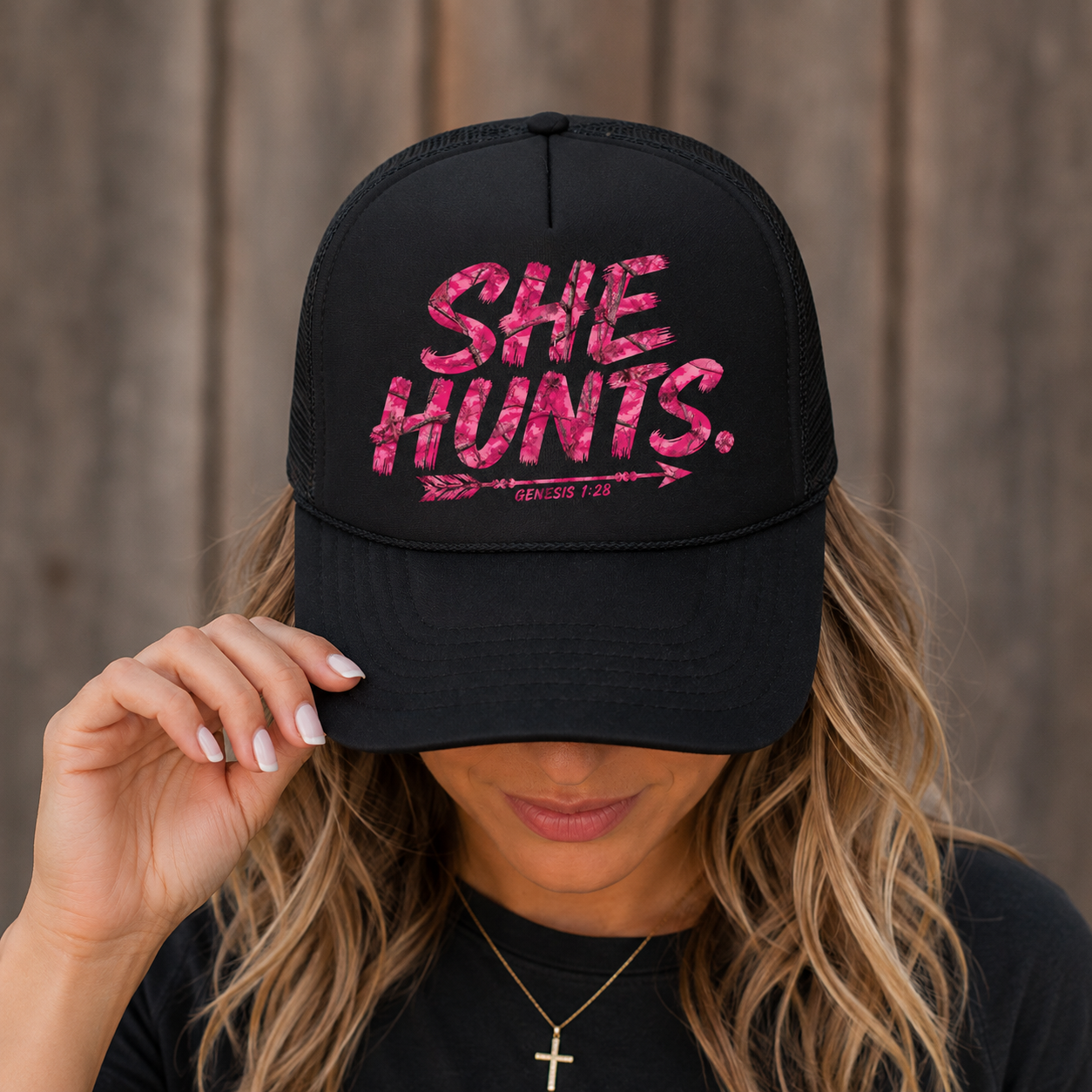 A woman wearing a black trucker hat with pink glitter-style text reading 'SHE HUNTS.' and smaller 'CINCUTS 128' text, photographed against a rustic wooden background.