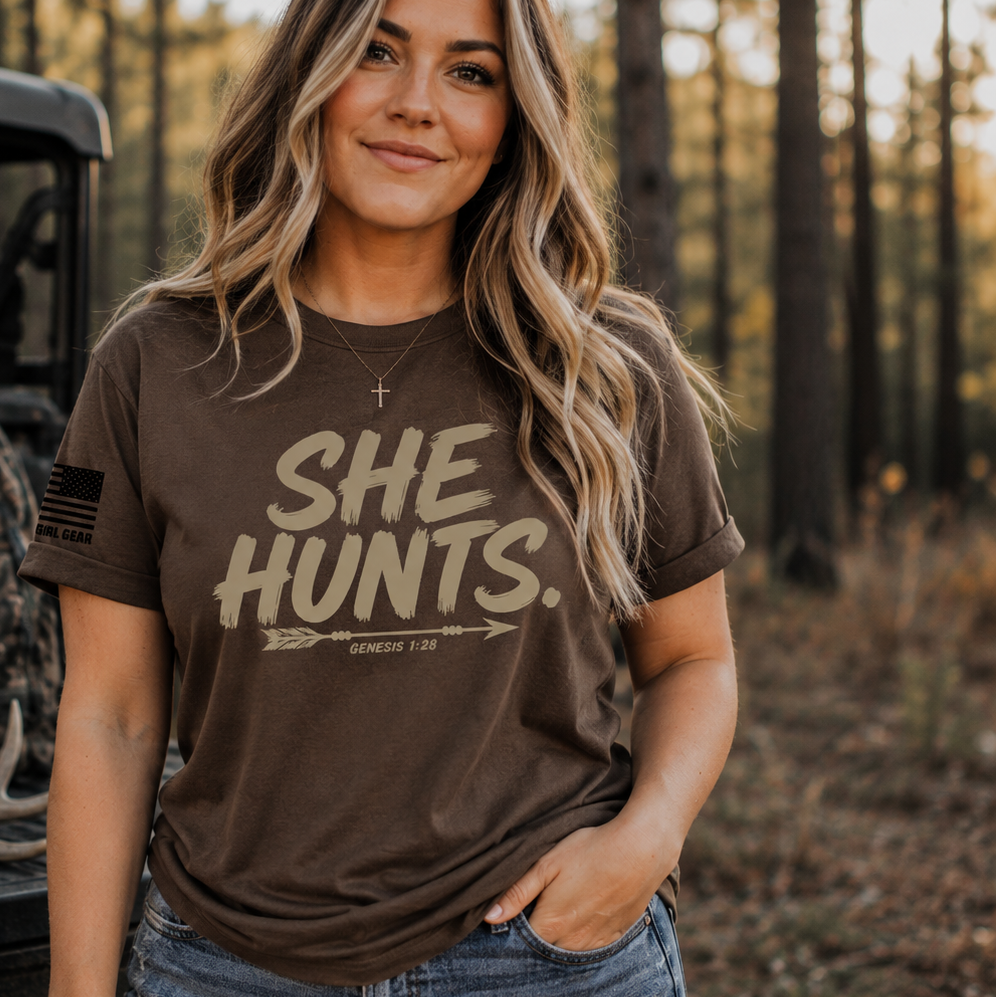 A woman wearing a brown graphic t-shirt with the text 'SHE HUNTS.' stands in a wooded outdoor setting next to a utility vehicle.