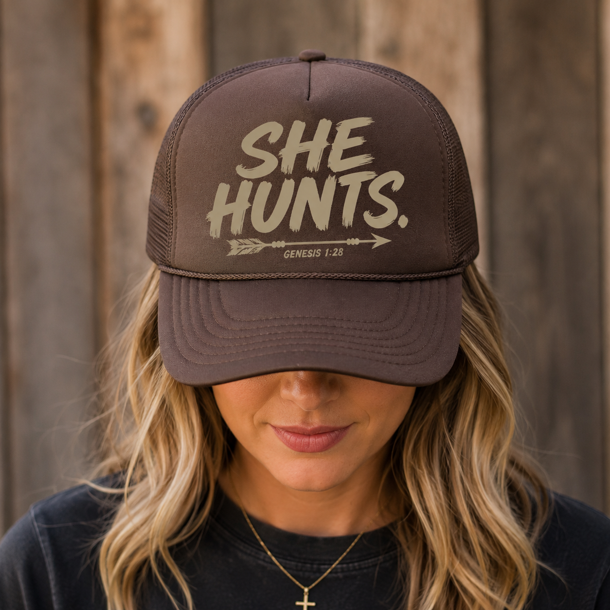 A woman wearing a dark brown trucker hat with the phrase 'SHE HUNTS.' and 'GENESIS 1:26' printed on the front, photographed against a rustic wooden background.