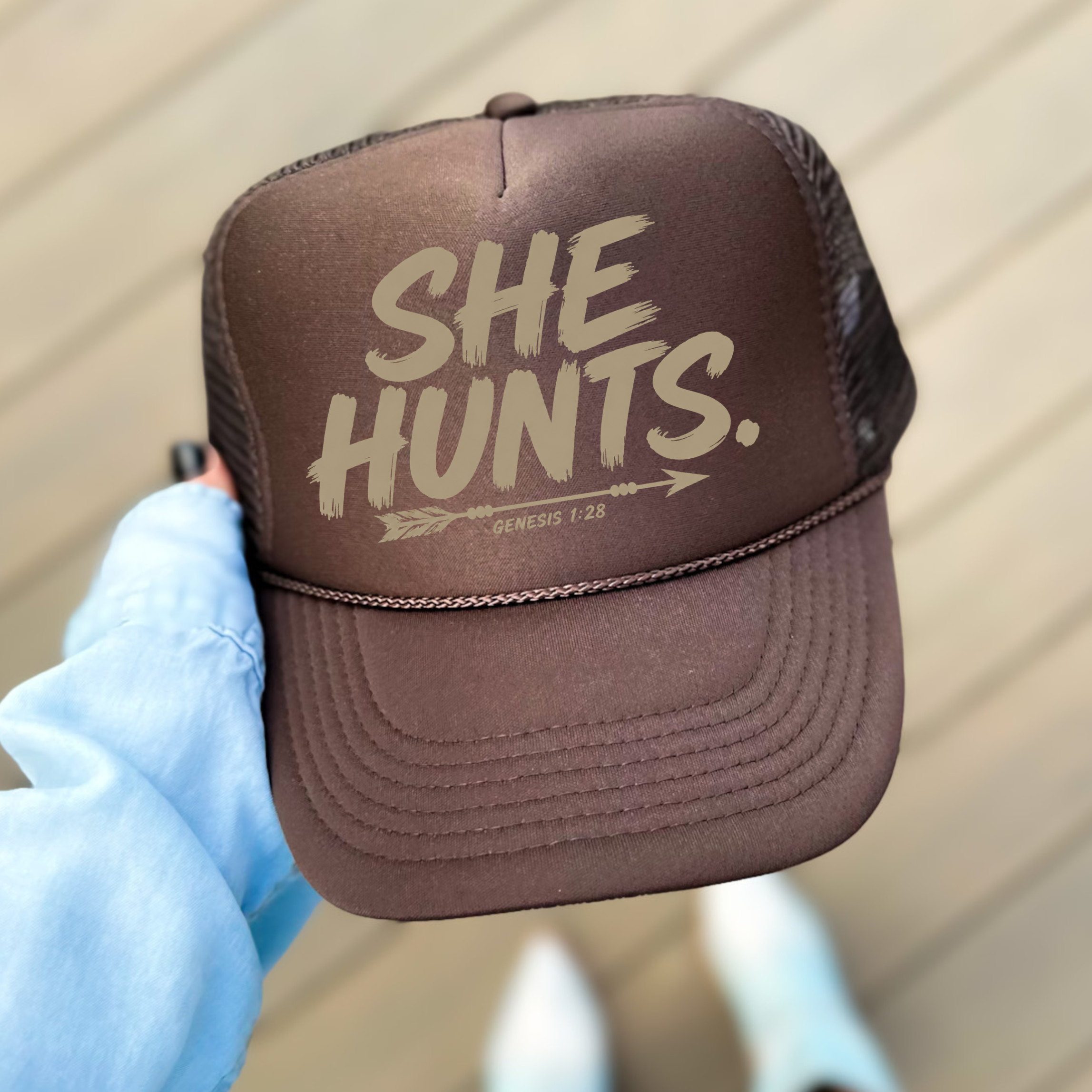 A person holds a brown trucker hat with the phrase 'SHE HUNTS.' printed in beige on the front panel.