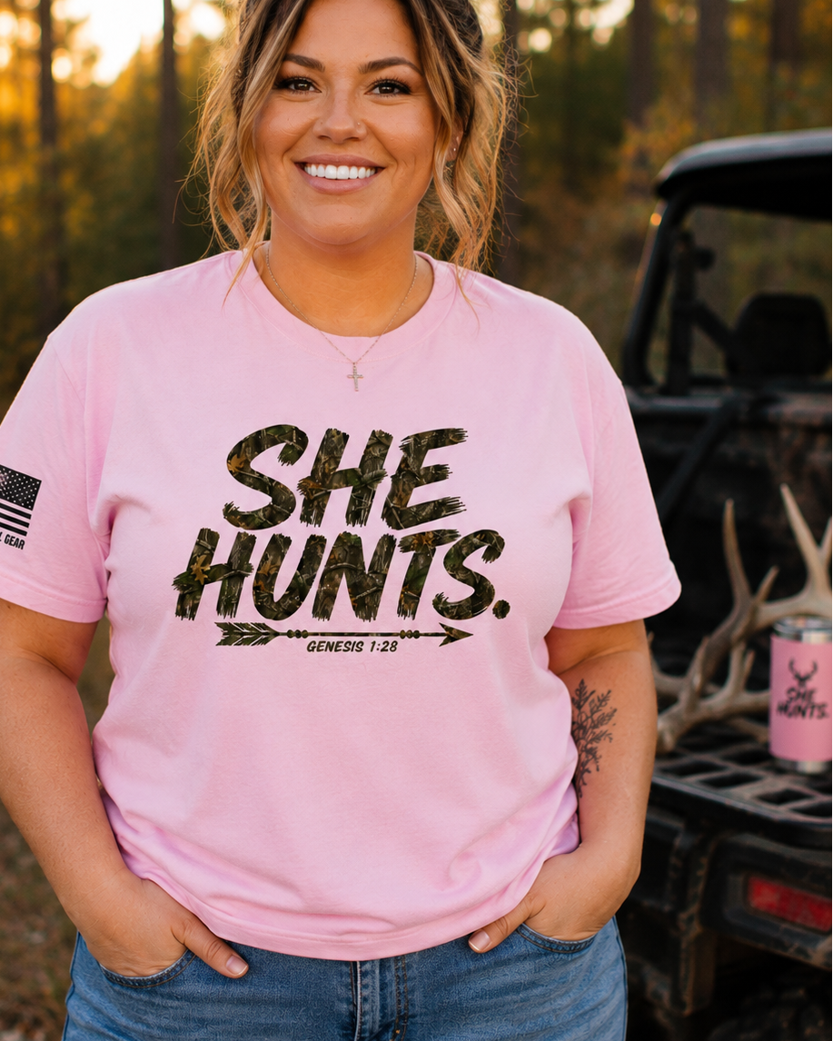 A woman wearing a light pink graphic t-shirt with the text 'SHE HUNTS.' in distressed black lettering, standing outdoors in a wooded setting next to a vehicle.