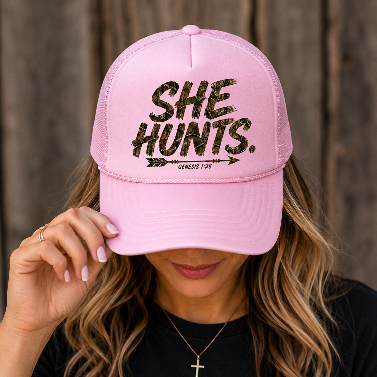 A woman wearing a light pink trucker hat with the text 'SHE HUNTS. GENESIS 1:26' in black and brown camouflage lettering, holding the brim with her hand against a rustic wooden background.