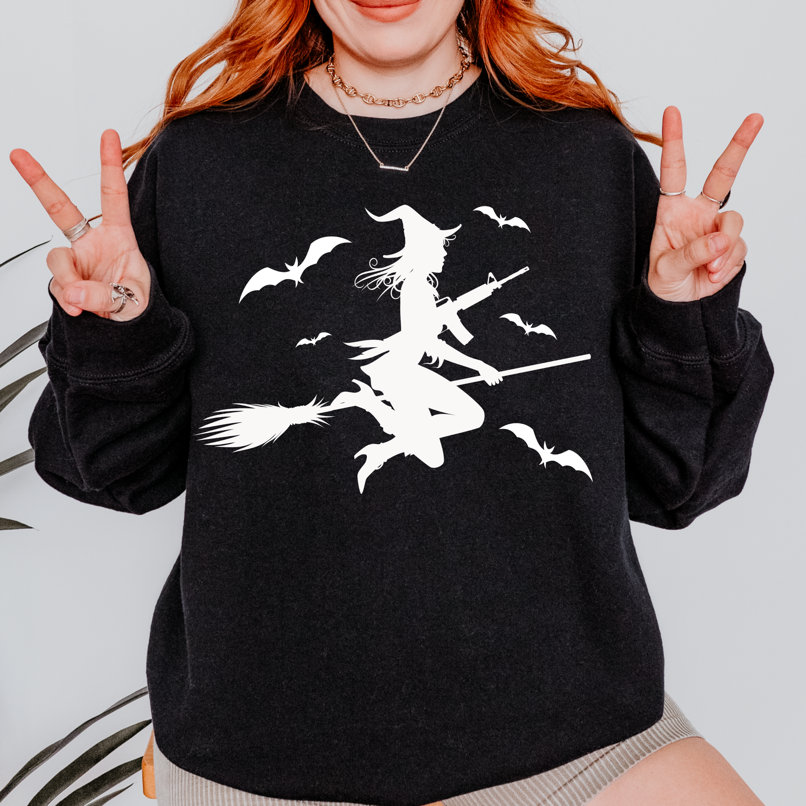 A woman wearing a black crewneck sweatshirt with a white graphic of a witch riding a broomstick with flying bats. She is posing with peace signs against a light gray background.